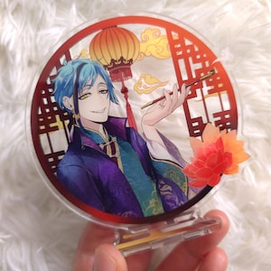 Puede incluir: Un soporte redondo de acrílico con una ilustración colorida de un hombre con cabello azul y una linterna roja. El hombre lleva una túnica morada y dorada y sostiene una pipa roja y dorada. El fondo es un diseño de estilo chino rojo y dorado.
