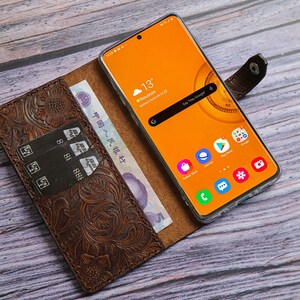 May include: A brown leather phone case with an embossed floral design, holding a smartphone with an orange screen displaying the weather in Houston. The case also holds credit cards and Chinese currency.