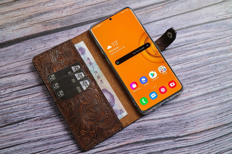 May include: A brown leather phone case with an embossed floral design, holding a smartphone with an orange screen displaying the weather in Houston. The case also holds credit cards and Chinese currency.