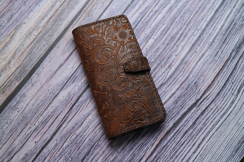 May include: A brown leather phone case with an embossed floral design. The case has a magnetic closure and a detailed pattern of flowers and leaves. The case is made of a rich, dark brown leather.