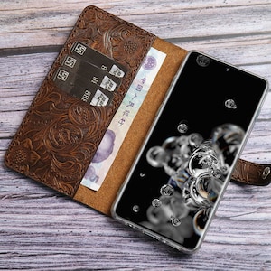 May include: Brown leather phone case with an embossed floral design. The case is open, revealing a smartphone with a black screen and a few credit cards. A bill is visible inside the case.