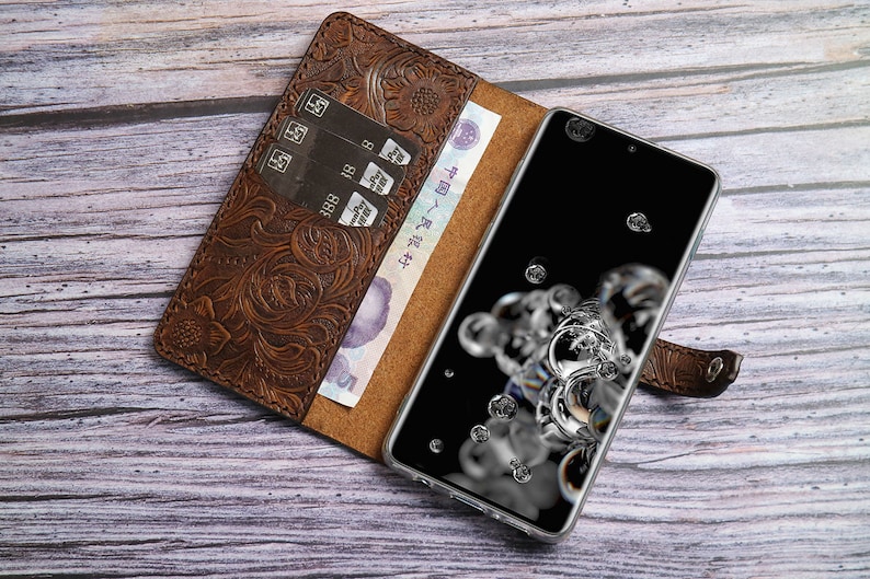 May include: Brown leather phone case with an embossed floral design. The case is open, revealing a smartphone with a black screen and a few credit cards. A bill is visible inside the case.