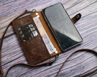 May include: A brown leather phone case with a floral embossed design, holding a smartphone, credit cards, and currency. The case has a detachable strap and silver-tone hardware.
