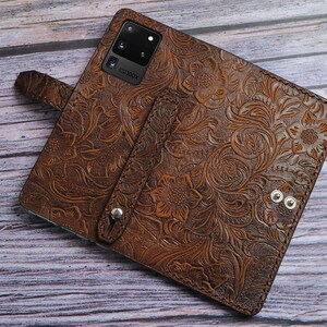 May include: A brown leather phone case featuring an intricate embossed floral pattern, a vertical strap with a silver metal snap button, and a rectangular cutout for a multi-lens camera system with the text 100X printed near the lens.