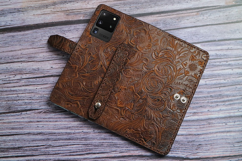 May include: A brown leather phone case featuring an intricate embossed floral pattern, a vertical strap with a silver metal snap button, and a rectangular cutout for a multi-lens camera system with the text 100X printed near the lens.