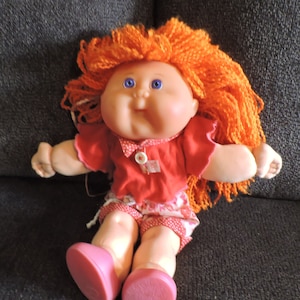 May include: A Cabbage Patch Kid doll with bright orange yarn hair, wearing a red shirt with white polka dots, pink and white polka dot shorts, and pink shoes.
