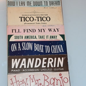 Op de afbeelding: Een stapel vintage bladmuziek met populaire liedjes uit de jaren 1940 en 1950, waaronder "When You're a Long, Long Way From Home", "Two in Love", "Now I Lay Me Down to Dream", "Tico-Tico", "I'll Find My Way", "South America, Take It Away", "On a Slow Boat to China", "Wanderin'", "Hey, Mr. Banjo" en "Dreamy Melody".