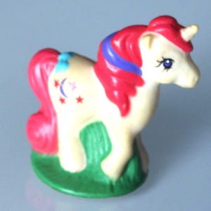 May include: A small, cream-coloured unicorn figurine with a red mane and tail, a blue stripe in its hair, and a small horn. The unicorn has a crescent moon and star design on its flank and stands on a green base.