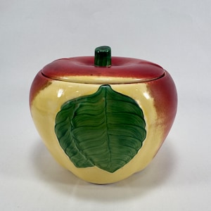 May include: A ceramic apple-shaped container with a lid. The top is red, transitioning to yellow on the sides. A large green leaf design is painted on the front. The stem is a small, dark green cylinder. This decorative piece is likely for kitchen or home decor.