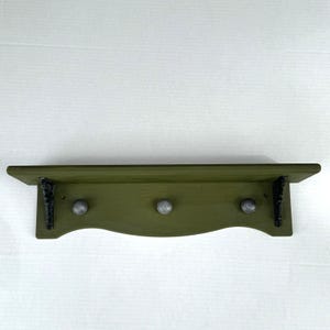 May include: A wall-mounted olive green wooden shelf featuring a flat top surface and a curved lower edge. The unit includes three round silver-toned knobs for hanging items and is supported by two ornate black metal brackets on the sides.
