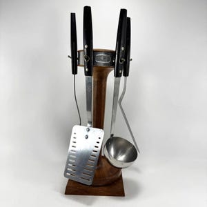 May include: A vintage kitchen utensil set with black handles and stainless steel tools. The set includes a spatula, ladle, and other tools, displayed on a wooden stand. The stand is a dark brown color.