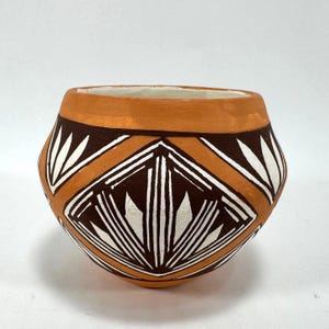 May include: A rounded terracotta pot featuring a wide, smooth orange rim and a body decorated with dark brown and white geometric patterns. The design consists of repeating diamond shapes filled with sharp, radiating white lines on a brown background.