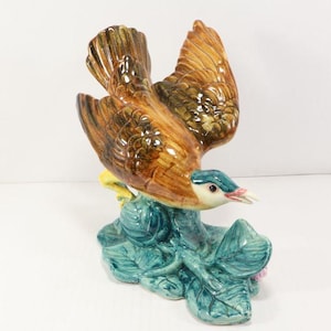 May include: A ceramic bird figurine with outstretched wings, featuring brown, tan, and teal colors. The bird has a teal head and is perched on a teal base with green leaves and a pink flower.