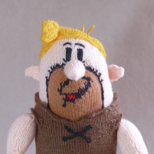 Può includere: Un giocattolo di peluche lavorato a maglia di un uomo delle caverne dei cartoni animati con un perizoma marrone, pelle rosa, capelli gialli e baffi e barba neri.