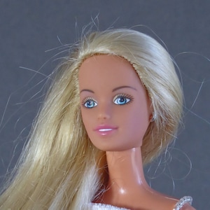 May include: A blonde doll with blue eyes wearing a white sleeveless top. The doll has a long, flowing hairstyle.