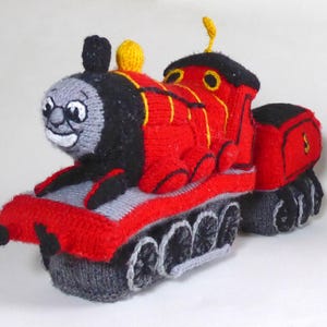 Op de afbeelding: Een rode en zwarte gebreide speelgoedtrein met een glimlachend gezicht. De trein heeft een gele fluit en zwarte wielen.