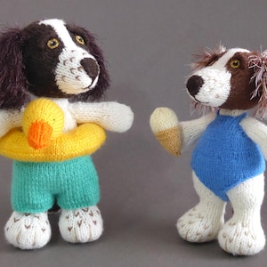 Puede incluir: Dos figuras de perros de punto. Uno lleva pantalones cortos turquesa y un salvavidas amarillo con un pato naranja. El otro perro lleva un bañador azul y sostiene un helado de punto. Ambos tienen pelaje blanco y marrón.