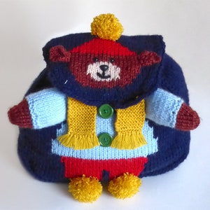 May include: A blue knitted backpack with a brown bear face on the front. The bear is wearing a red hat, a yellow scarf, and a blue sweater. The backpack has two yellow pom-poms on the bottom.