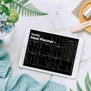 May include: A white tablet displaying a black and white "Weekly Meal Planner" layout. The tablet is surrounded by a light blue knitted scarf, flowers in a glass vase, and a wooden tray with a watch and accessories. Fern leaves and a notebook are also visible.