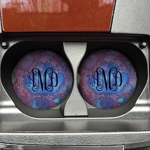 May include: Two round car coasters with a blue, pink, and white watercolor design and glitter accents. The coasters have the monogram "AMD" in black.