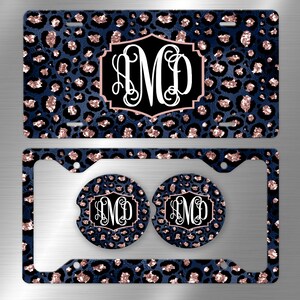 May include: A set of two car accessories featuring a blue and rose gold leopard print design. The set includes a number plate frame and two cup coasters. The number plate frame has a monogrammed initial "M" in the centre. The coasters also feature the same monogrammed initial.