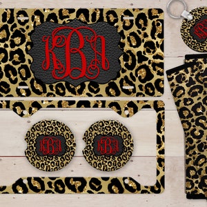 May include: A set of car accessories featuring a leopard print design with gold glitter accents. The set includes a number plate frame, two cup holders, a key ring, and a seatbelt cover. The number plate frame and cup holders feature a monogram in red.