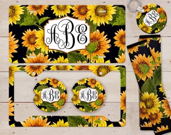 May include: A set of car accessories featuring a sunflower design. The set includes a license plate frame, two coasters, a key chain, and a seatbelt strap. The accessories are all black with a sunflower pattern and the initials "HBE" in a white circle.