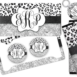 May include: A set of car accessories featuring a black and white leopard print design with silver glitter accents. The set includes a license plate frame, two cup holders, a key chain, and two seat belt covers. The monogram "SKP" is featured on each item.
