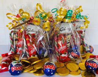 Puede incluir: Varias cestas de regalo envueltas en celofán llenas de barras de chocolate, bastones de caramelo y monedas de chocolate doradas. Las cestas están atadas con cintas doradas y verdes. Se ven varias marcas de chocolate.