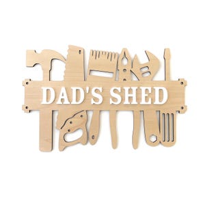 May include: Wooden sign with the words "Dad's Shed" in white lettering. The sign is decorated with cutouts of various tools, including a hammer, saw, wrench, screwdriver, and spirit level.