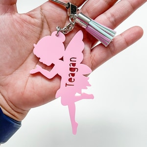 May include: Pink fairy keyring with a silver charm and a pink and white tassel. The fairy is holding a flower and the name Teagan is engraved on the keyring.