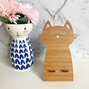 May include: A wooden phone stand shaped like a cat with closed eyes. The stand is made of light brown bamboo and has two slots for holding a phone.