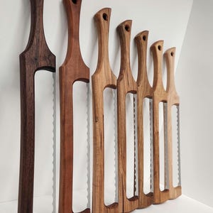 May include: A collection of wooden bread knives with serrated blades. The handles vary in color from dark brown to light tan. Each knife has a hole at the top of the handle. The knives are displayed against a white background.