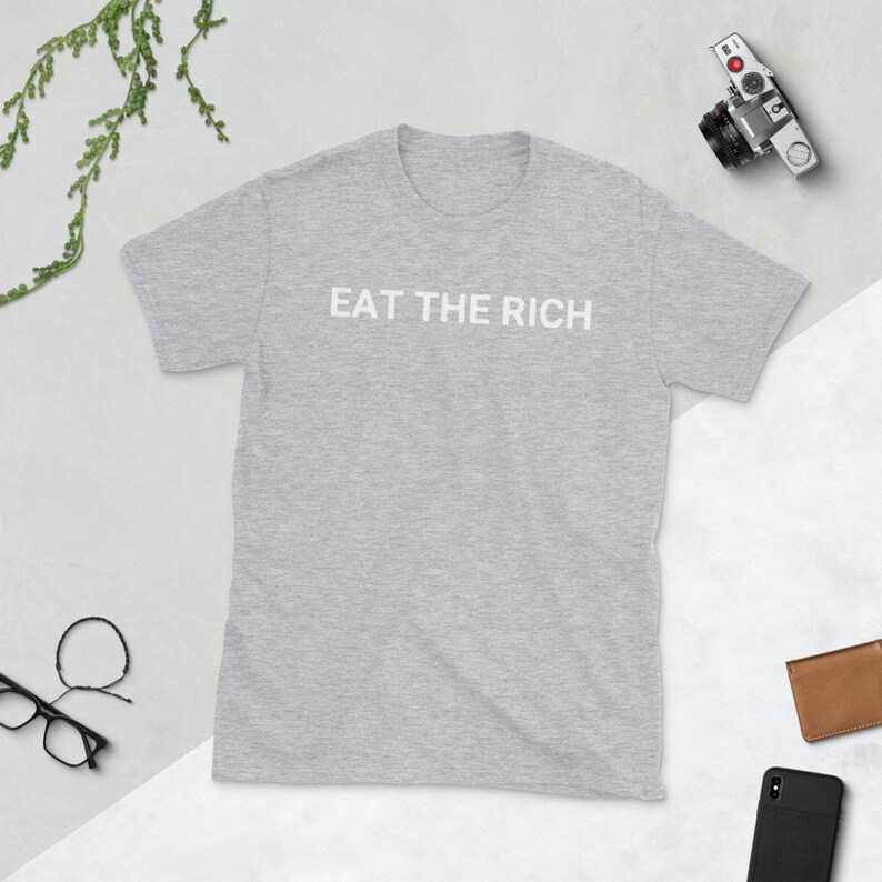 May include: A light gray t-shirt with the text "EAT THE RICH" printed in white across the chest. The shirt is short-sleeved and has a crew neck. A camera, wallet, phone, and glasses are next to the shirt.