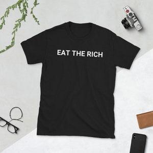 May include: Black t-shirt with the white text "EAT THE RICH" across the chest. The shirt is short-sleeved and appears to be made of a soft material. A camera, glasses, and a wallet are visible in the background.