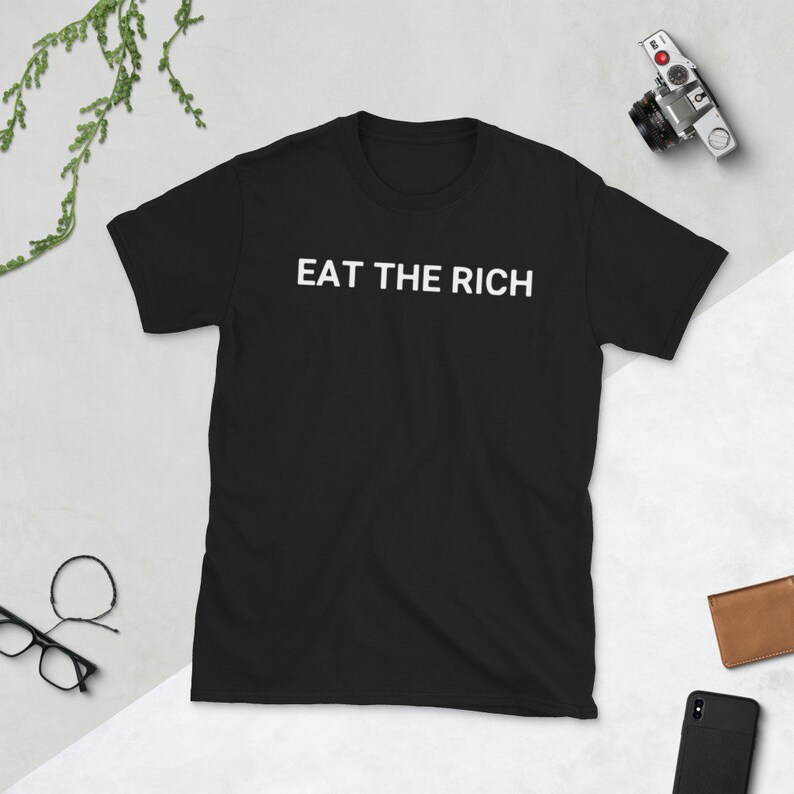 May include: Black t-shirt with the white text "EAT THE RICH" across the chest. The shirt is short-sleeved and appears to be made of a soft material. A camera, glasses, and a wallet are visible in the background.