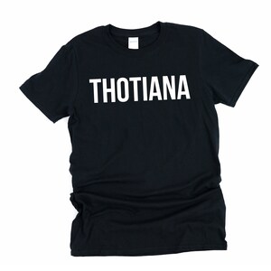 May include: Black t-shirt with the word "THOTIANA" printed in large, white capital letters across the chest. The shirt is a basic crew neck style, likely made of cotton or a similar fabric, and is displayed on a white background.
