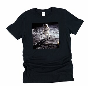 May include: Black t-shirt featuring a graphic of an astronaut on the moon with the text "NEVADA 1969" in white. The image shows the astronaut's shadow and the lunar surface. The shirt is a crew neck style.