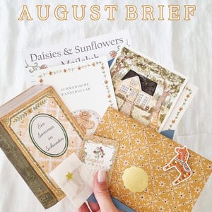 Könnte beinhalten: Ein Schreibwaren-Set mit der Aufschrift AUGUST BRIEF, bestehend aus einem Notizblock Daisies & Sunflowers, einer Karte mit Haus-Illustration, einem Buch mit dem Titel Ein Sommer in Schweden, einem kleinen August-Anhänger und einem gelben Umschlag.