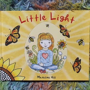 May include: A yellow book with the title "Little Light" in black text. The cover features a girl with long blonde hair sitting in a lotus position with a red heart on her shirt. The girl is surrounded by butterflies, bees, and flowers.