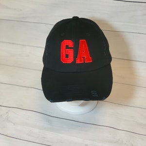 May include: Black baseball cap with distressed detailing. The cap features the red embroidered letters "GA" on the front. The hat is displayed on a white stand against a white wooden background.