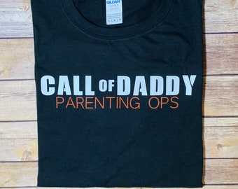May include: Black t-shirt with the text "CALL OF DADDY" in white and "PARENTING OPS" in orange. The shirt is folded and displayed on a wooden surface. The brand label "GILDAN" is visible.