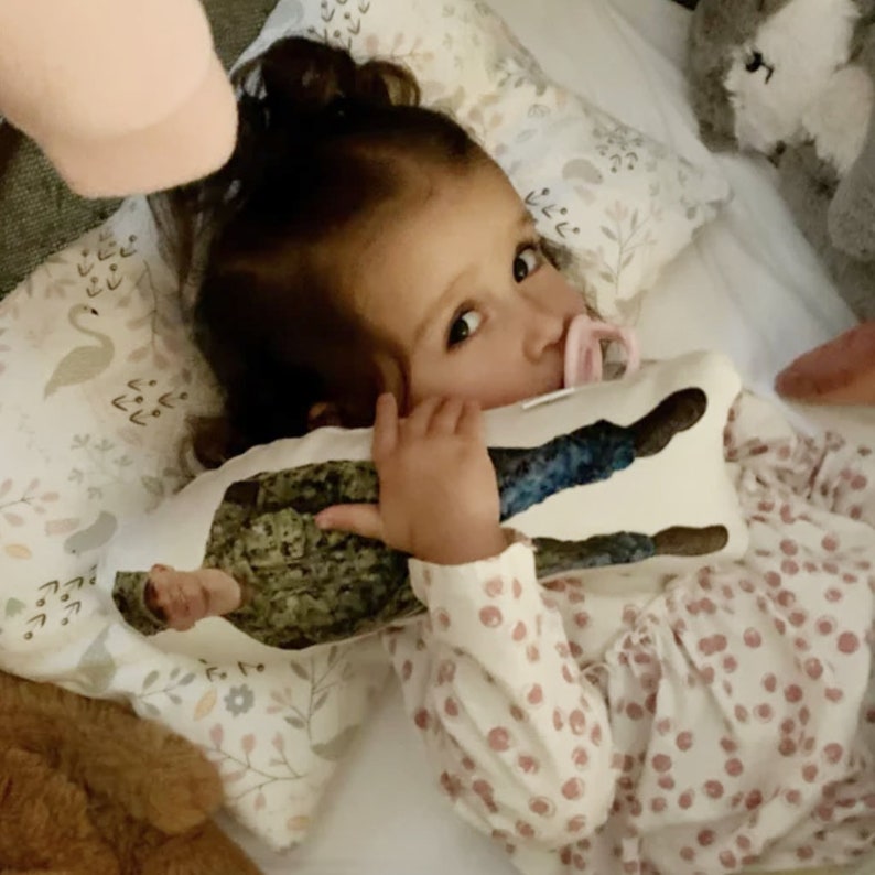 Pu&ograve; includere: Una bambina con i capelli castani &egrave; sdraiata a letto con un cuscino bianco con un motivo floreale. La bambina tiene in mano un cuscino con un motivo mimetico e un'immagine di una persona in uniforme militare. La bambina indossa una camicia bianca con pois rosa.
