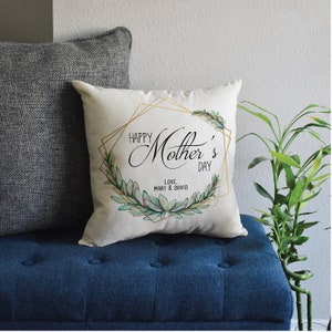 May include: A white pillow with a gold geometric frame and green leafy design. The text "Happy Mother's Day" is written in black cursive font. The text "Love, Mary & David" is written in black font below the main text.
