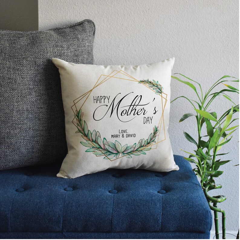 May include: A white pillow with a gold geometric frame and green leafy design. The text "Happy Mother's Day" is written in black cursive font. The text "Love, Mary & David" is written in black font below the main text.