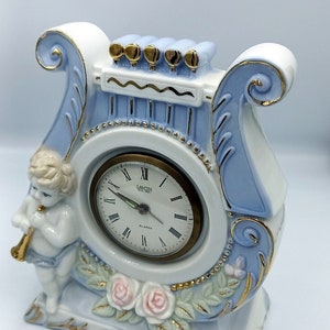 Pode incluir: Um relógio de porcelana branco e azul em forma de lira com um querubim tocando flauta. O mostrador do relógio é branco com algarismos romanos pretos e ponteiros dourados. O relógio tem uma moldura dourada e detalhes florais.