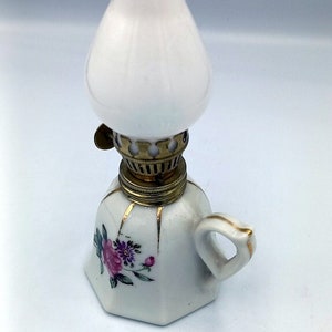 Op de afbeelding: Een kleine witte porseleinen olielamp met een bloemenmotief en een gouden rand. De lamp heeft een witte glazen kap en een messing lont houder.