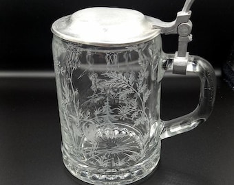 May include: A clear glass beer stein with an etched design of a deer in a forest. The stein has a silver lid with a hinged handle.
