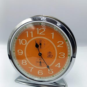 May include: A chrome-finished, round analog clock with an orange face and white numerals. The clock features a smaller dial and the brand name "HOFEA". The clock has a curved base and a glass cover.