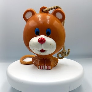 May include: A brown plastic toy bear with blue eyes, a red nose, and a white face. The bear has a string attached to its head and is sitting on a white surface.